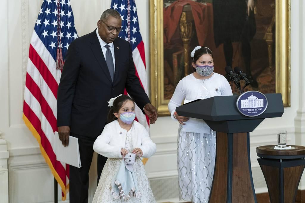 Gabby Rodriguez (right) and her sister, Eva (center), are assisted to the stage in the East Room of the White House by Defense Secretary Lloyd Austin (left) during a ceremony at the White House honoring children in militar Kids of wounded veterans to get support, attention under new â€˜hidden helpersâ€™ campaign